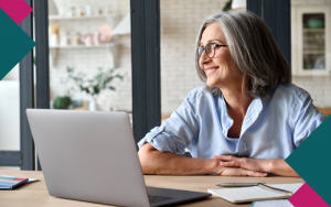 Woman with glasses and gray hair looks out window while using laptop to work on her small business marketing goals